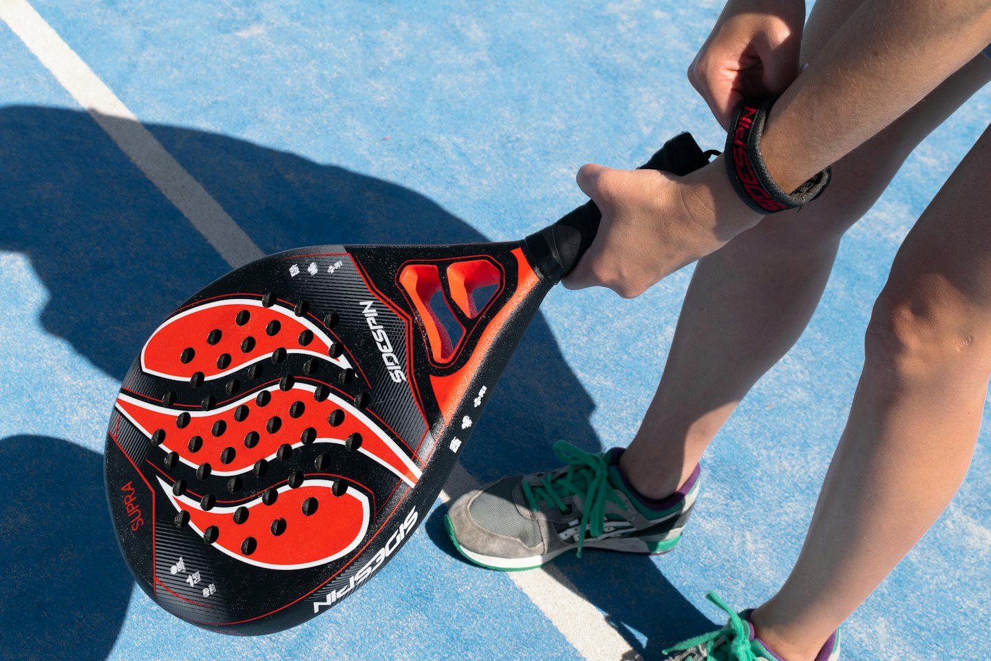 Mani che aggiustano il laccio della racchetta su un campo da padel blu.
